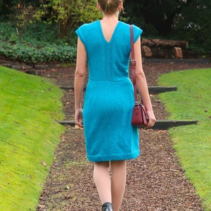 May include: A woman walks down a path wearing a turquoise blue sleeveless dress and black heeled boots. She is carrying a small burgundy handbag.