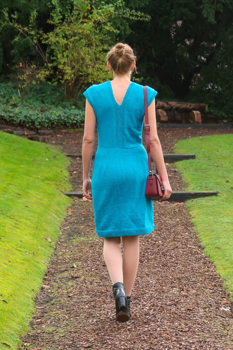 May include: A woman walks down a path wearing a turquoise blue sleeveless dress and black heeled boots. She is carrying a small burgundy handbag.