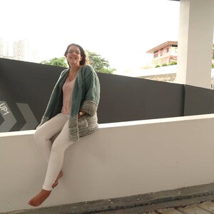 May include: A woman wearing a teal and white knitted cardigan with a pink tank top underneath and white pants. She is sitting on a white wall with a black and white arrow sign that says "UP1" on it.