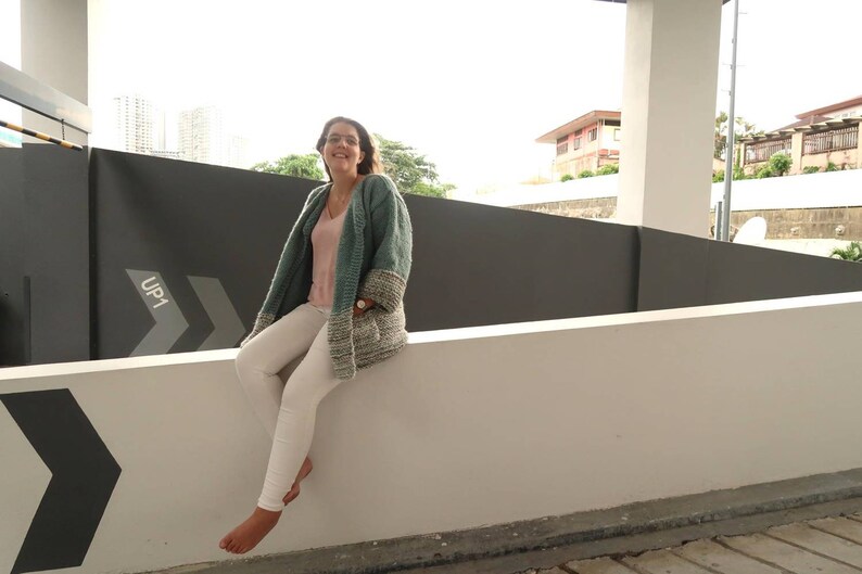 May include: A woman wearing a teal and white knitted cardigan with a pink tank top underneath and white pants. She is sitting on a white wall with a black and white arrow sign that says "UP1" on it.