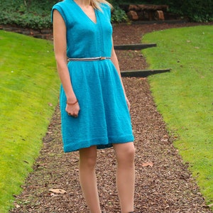 May include: A woman wearing a turquoise blue knitted dress with a brown belt and black ankle boots. She is standing on a path with brown mulch and green grass.