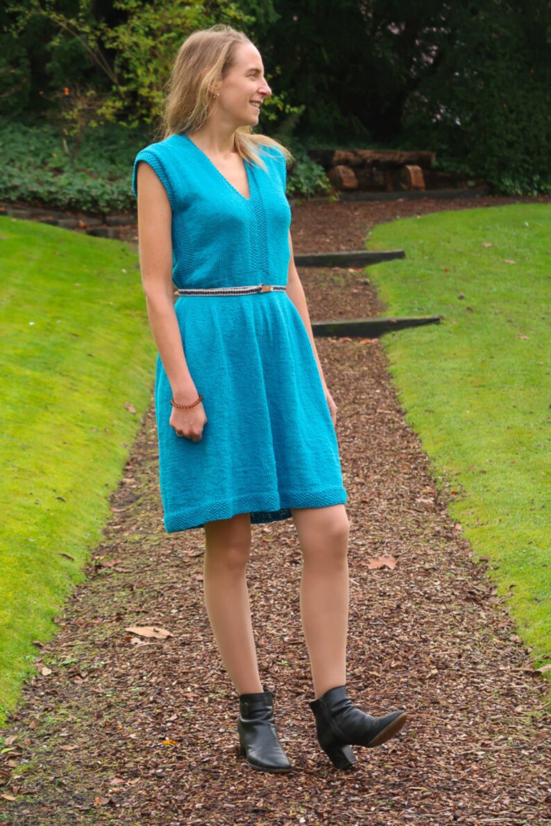 May include: A woman wearing a turquoise blue knitted dress with a brown belt and black ankle boots. She is standing on a path with brown mulch and green grass.