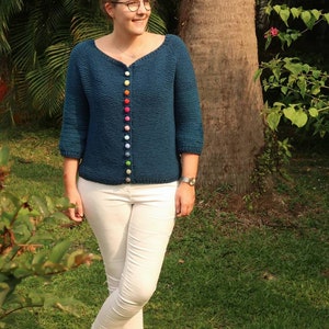 K&ouml;nnte beinhalten: Ein blaugr&uuml;ner Cardigan mit einem Regenbogen aus bunten Kn&ouml;pfen. Der Cardigan ist gestrickt und hat einen V-Ausschnitt. Die Person, die den Cardigan tr&auml;gt, steht in einem grasbewachsenen Bereich.