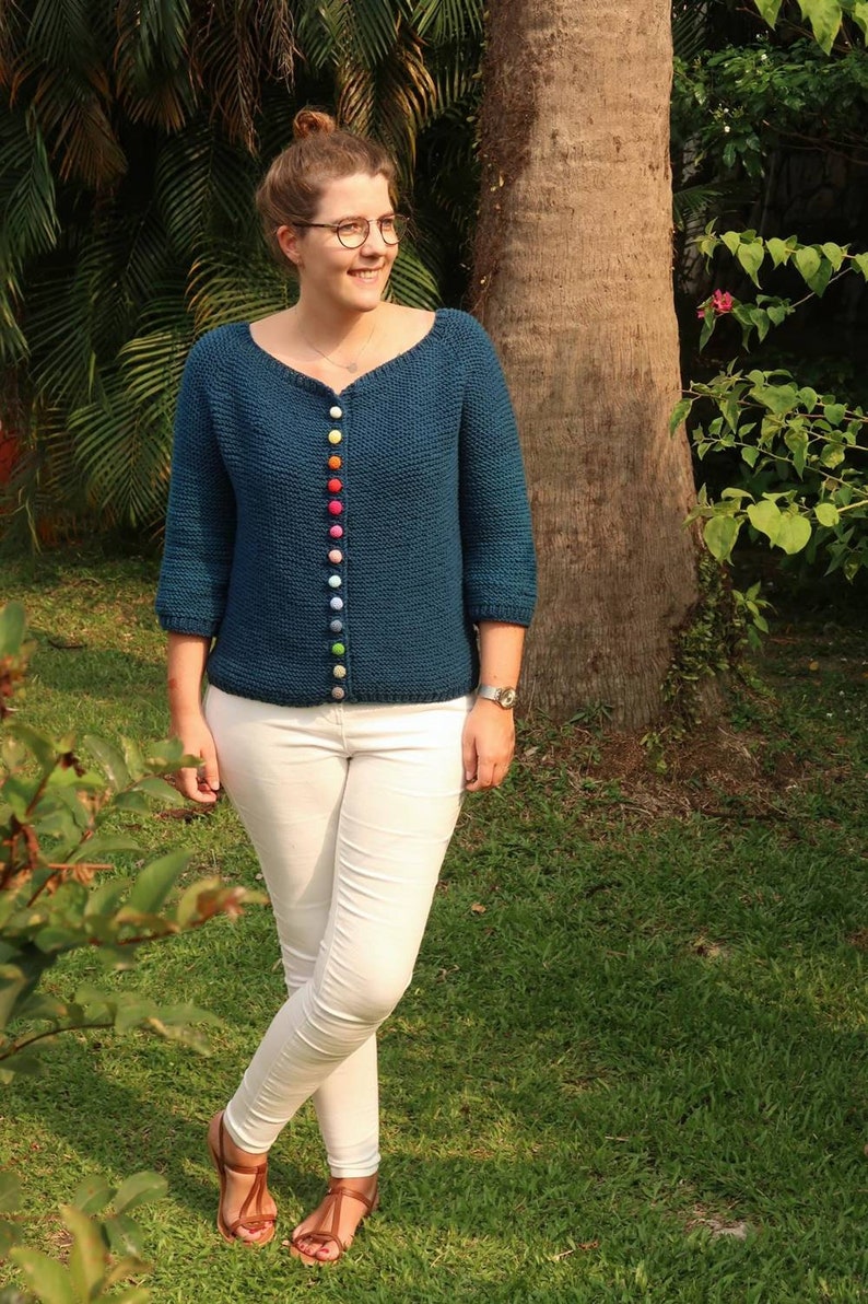 K&ouml;nnte beinhalten: Ein blaugr&uuml;ner Cardigan mit einem Regenbogen aus bunten Kn&ouml;pfen. Der Cardigan ist gestrickt und hat einen V-Ausschnitt. Die Person, die den Cardigan tr&auml;gt, steht in einem grasbewachsenen Bereich.