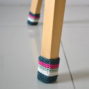 Puede incluir: Primer plano de una pata de silla de madera con una cubierta de punto de rayas azul verdoso, blanco y rosa. La cubierta está diseñada para proteger los pisos de los arañazos.
