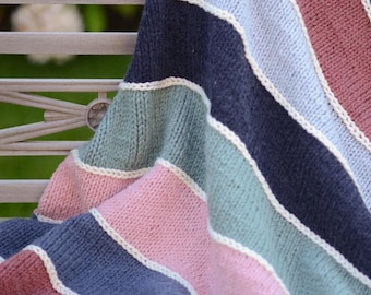 May include: A striped knitted throw blanket in shades of pink, blue, green, and brown with white accents. The blanket is folded and draped over a wooden bench.