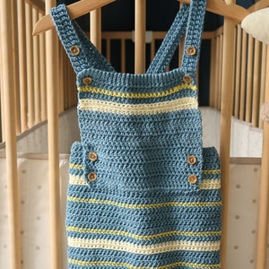 May include: A blue and white striped crocheted baby overall with wooden buttons. The overall is hanging on a wooden hanger.