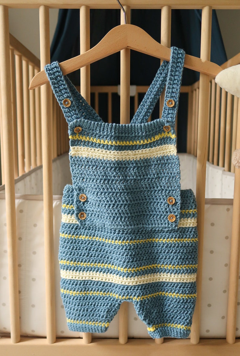 May include: A blue and white striped crocheted baby overall with wooden buttons. The overall is hanging on a wooden hanger.