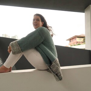 May include: A woman wearing a teal knitted cardigan with white and grey trim sits on a white wall. She is wearing white pants and has her feet on the wall. The sign "UP1" is visible in the background.