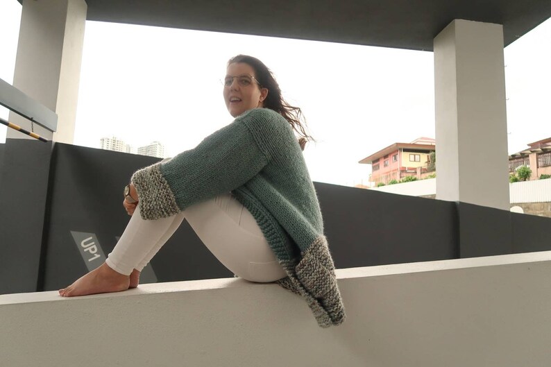 May include: A woman wearing a teal knitted cardigan with white and grey trim sits on a white wall. She is wearing white pants and has her feet on the wall. The sign "UP1" is visible in the background.