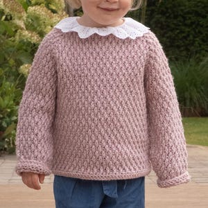 Op de afbeelding: Een roze gebreide trui met een witte kantkraag. De trui heeft een textuur met een ruitpatroon.