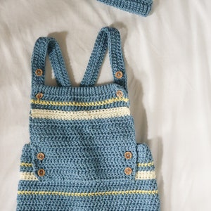 May include: A blue and white striped crocheted baby romper with yellow accents and wooden buttons. The romper has straps and a bib-like top.