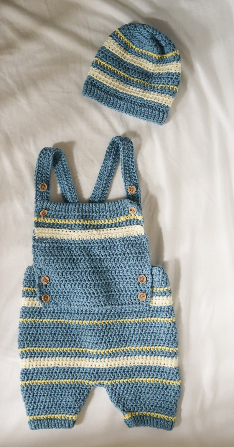 May include: A blue and white striped crocheted baby romper with yellow accents and wooden buttons. The romper has straps and a bib-like top.