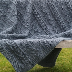 May include: A folded, knitted blanket in a muted blue-grey hue. The blanket features a textured, diagonal pattern with raised stitches and a thick, cosy appearance. It is draped over a wooden surface, with a grassy background.