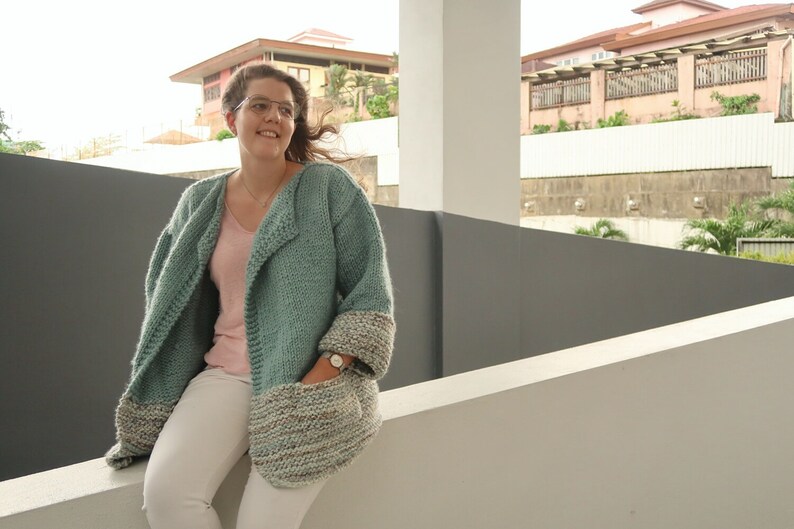 May include: A teal blue knitted cardigan with a shawl collar and pockets. The cardigan has a contrasting white and brown knit trim on the sleeves and pockets.
