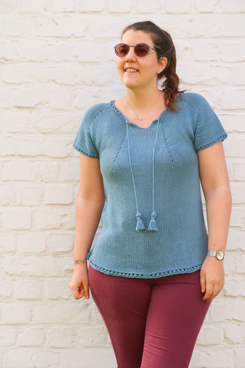 Pu&ograve; includere: Un top in maglia azzurra con maniche corte e scollo a V. Il top presenta una coulisse con nappe ed &egrave; abbinato a pantaloni bordeaux. Lo sfondo &egrave; un muro di mattoni bianchi.