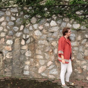 Peut inclure: Une femme portant un cardigan en crochet rouge avec des bordures blanches, un pantalon blanc et des chaussures grises se tient devant un mur en pierre avec des vignes vertes.
