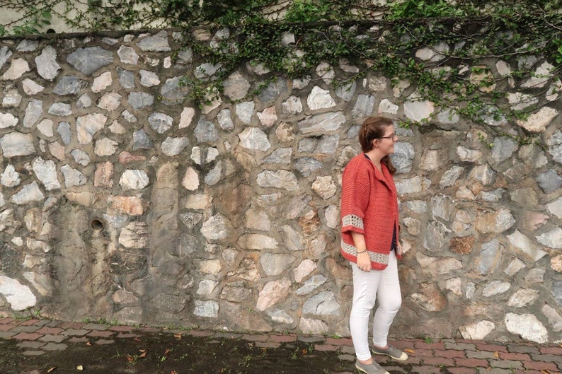 Peut inclure: Une femme portant un cardigan en crochet rouge avec des bordures blanches, un pantalon blanc et des chaussures grises se tient devant un mur en pierre avec des vignes vertes.