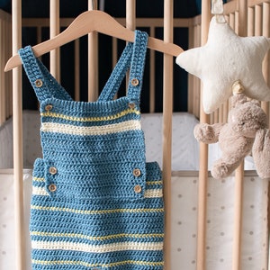 May include: A blue and white striped crocheted baby overall with wooden buttons hanging on a wooden hanger in a crib.