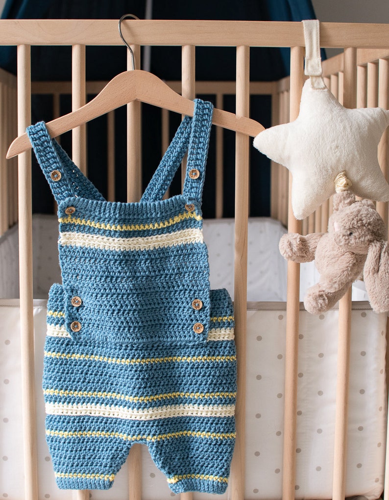 May include: A blue and white striped crocheted baby overall with wooden buttons hanging on a wooden hanger in a crib.