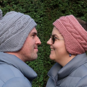 May include: A man and woman are seen wearing knitted winter accessories. The man wears a gray cable-knit hat with a pom-pom, and the woman wears a pink cable-knit headband. Both are wearing gray jackets, and the background is green.