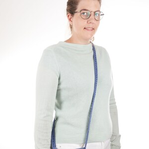 May include: A woman wearing a light green sweater and white pants is holding a blue crocheted crossbody bag with a wooden butterfly accent.