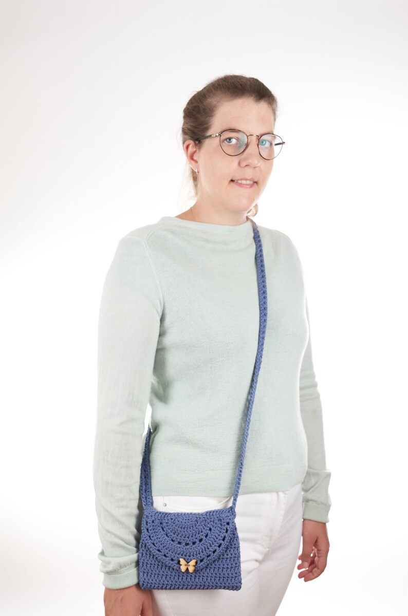 May include: A woman wearing a light green sweater and white pants is holding a blue crocheted crossbody bag with a wooden butterfly accent.