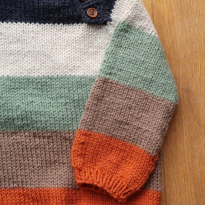 Op de afbeelding: Een gestreepte gebreide trui met een navyblauwe, witte, groene, bruine en oranje kleurstelling. De trui heeft een knoopsluiting.