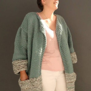 May include: A teal knitted cardigan with brown and white trim on the sleeves and pockets. The cardigan is open in the front and has a relaxed fit.