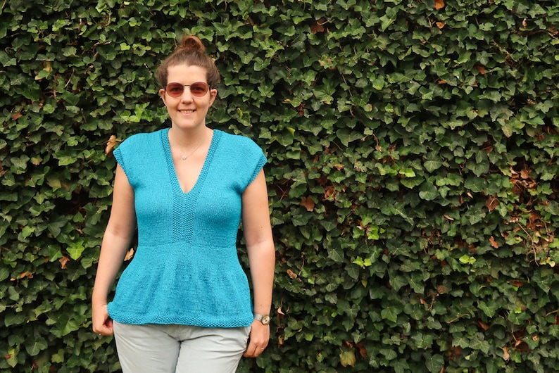 May include: A woman wearing a teal blue knitted top with a v-neck and short sleeves. She is standing in front of a green ivy wall.