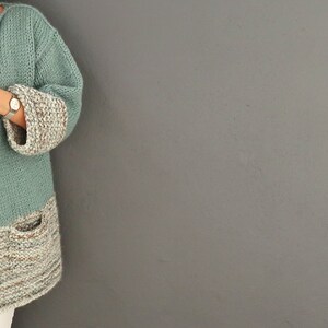 May include: A teal knitted cardigan with a brown and white striped bottom and cuffs. The cardigan has a pocket on the left side.