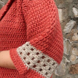 Peut inclure: Un cardigan en crochet de couleur rouge corail avec une bordure beige autour des manches. Le cardigan est r&eacute;alis&eacute; avec un point textur&eacute;.