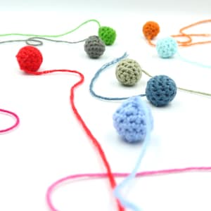 K&ouml;nnte beinhalten: Eine Sammlung bunter geh&auml;kelter Kugeln, die mit Garn aneinandergereiht sind. Die Kugeln sind in den Farben Rot, Rosa, Blau, Gr&uuml;n, Grau und Orange.