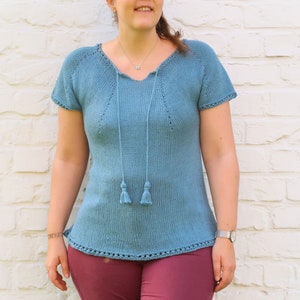 Pu&ograve; includere: Un top in maglia azzurra con maniche corte e scollo a V, con coulisse e nappe. Il top &egrave; abbinato a pantaloni bordeaux. La donna indossa occhiali da sole e un orologio.