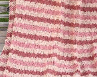 May include: A hand-knitted blanket with horizontal stripes in shades of pink, cream, and mauve. The blanket is made of thick yarn and has a textured appearance. The edges are finished with a solid mauve border.