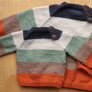 Op de afbeelding: Twee gestreepte gebreide truien in oranje, bruin, groen, wit en marineblauw. De truien hebben houten knopen op de schouders.