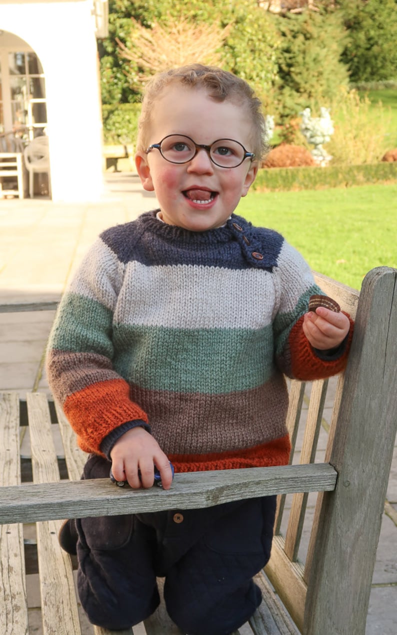 Op de afbeelding: Een jonge jongen draagt een gestreepte trui met oranje, bruin, groen en witte strepen. Hij draagt een blauwe spijkerbroek en heeft een speelse uitdrukking op zijn gezicht. Hij houdt een klein voorwerp in zijn hand.