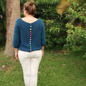 K&ouml;nnte beinhalten: Ein tealblauer Strickpullover mit einer Reihe bunter Kn&ouml;pfe auf dem R&uuml;cken.
