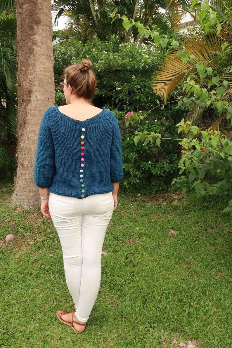 K&ouml;nnte beinhalten: Ein tealblauer Strickpullover mit einer Reihe bunter Kn&ouml;pfe auf dem R&uuml;cken.