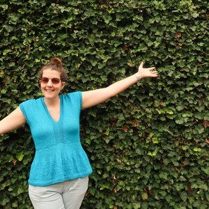 May include: A woman wearing a light blue, short-sleeved top with a V-neck and a ruffled hem, standing in front of a green ivy wall. She is smiling and has her arms outstretched.
