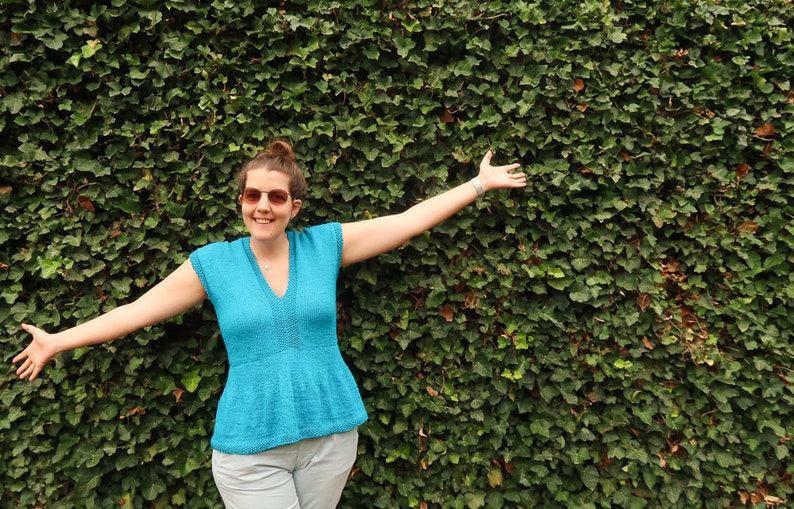 May include: A woman wearing a light blue, short-sleeved top with a V-neck and a ruffled hem, standing in front of a green ivy wall. She is smiling and has her arms outstretched.