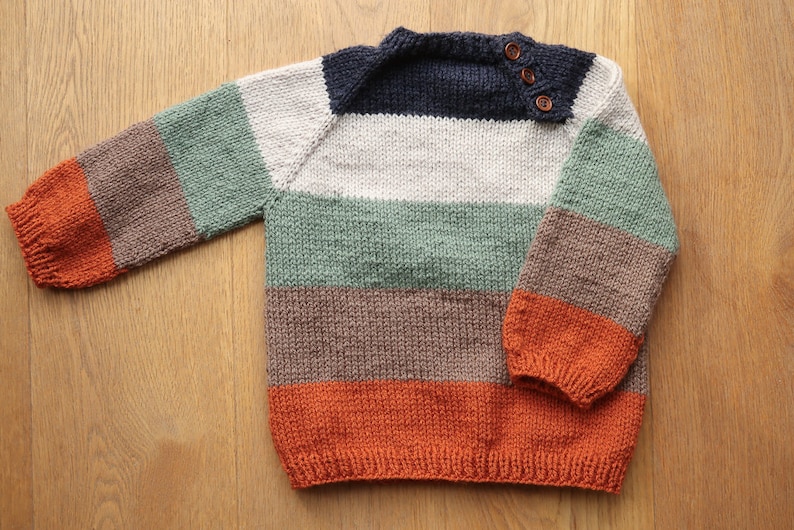 Op de afbeelding: Een gestreepte gebreide trui met lange mouwen en een ronde hals. De trui is in tinten oranje, bruin, groen, wit en donkerblauw. Er zitten drie bruine knopen op de schoudernaad.