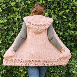 Könnte beinhalten: Eine rosa Strickweste mit Kapuze und Taschen. Die Weste hat einen dekorativen Besatz am unteren Rand.