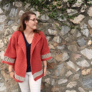 Peut inclure: Un cardigan en crochet rouge avec un col ch&acirc;le et des bordures en crochet blanc. Le cardigan est port&eacute; sur une chemise noire et un pantalon blanc.