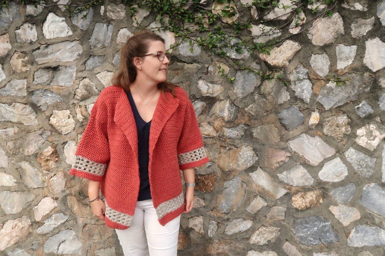 Peut inclure: Un cardigan en crochet rouge avec un col ch&acirc;le et des bordures en crochet blanc. Le cardigan est port&eacute; sur une chemise noire et un pantalon blanc.