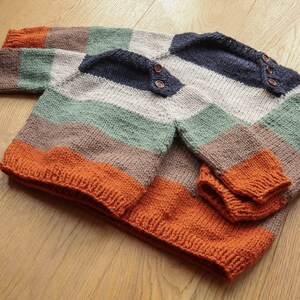 Op de afbeelding: Een gestreepte gebreide trui in tinten oranje, bruin, groen en blauw met houten knopen.