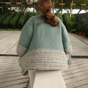May include: A light green knitted cardigan with a multi-colored bottom band. The cardigan is made of a soft, chunky yarn and has a relaxed fit.