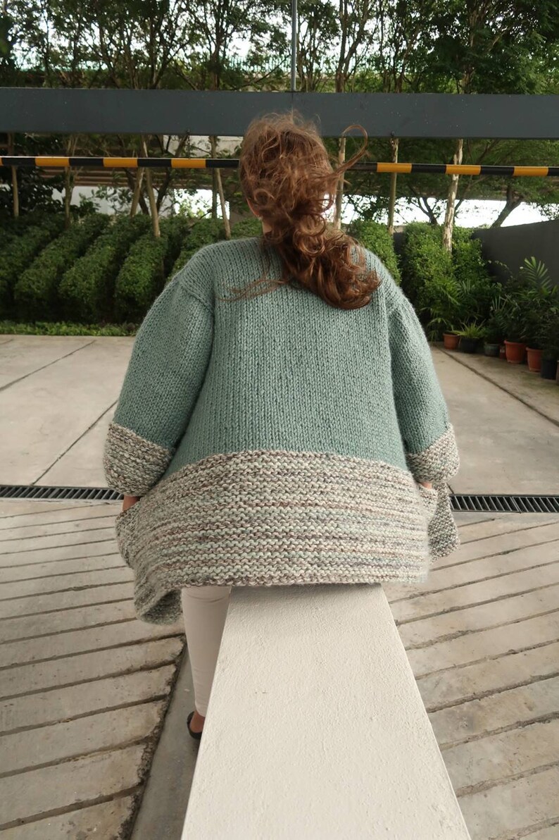 May include: A light green knitted cardigan with a multi-colored bottom band. The cardigan is made of a soft, chunky yarn and has a relaxed fit.