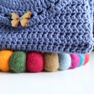 May include: A blue crocheted bag with a wooden butterfly button and a colorful felt pom-pom trim.