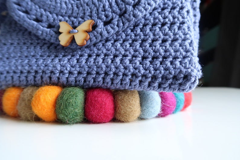 May include: A blue crocheted bag with a wooden butterfly button and a colorful felt pom-pom trim.